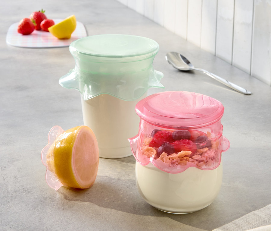 Na blacie kuchennym stoją dwa słoiki z jogurtem z różowymi i miętowymi pokrywkami oraz połówka cytryny z różową pokrywką.