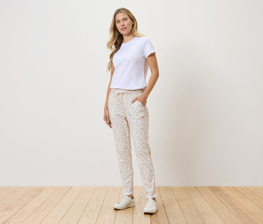 Kobieta stoi na tle ściany, ubrana w białą koszulkę, wzorzyste spodnie i białe tenisówki.