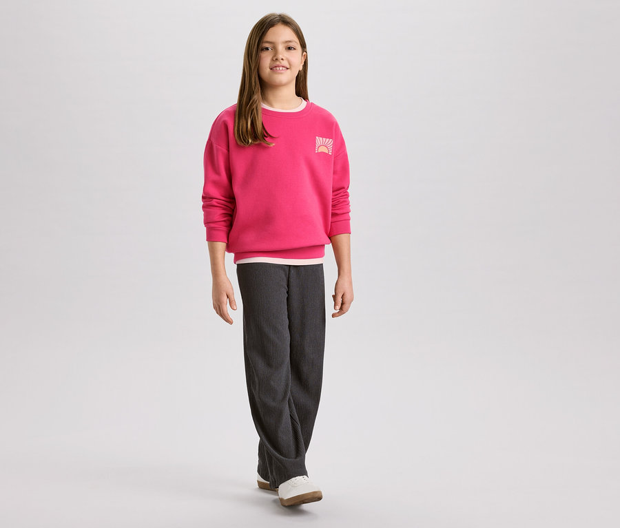 Dziewczyna w różowej bluzie z logo wschodu słońca i szarych sztruksowych spodniach, do tego białe buty.