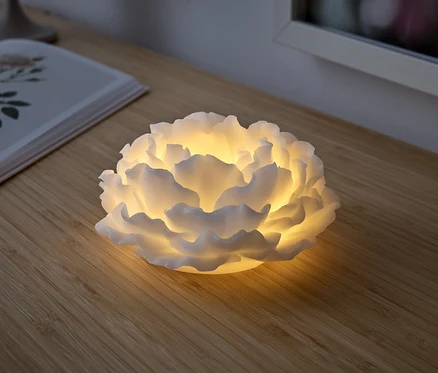 Biała lampa w kształcie kwiatu świeci na drewnianym stole. Obok leży otwarta książka.