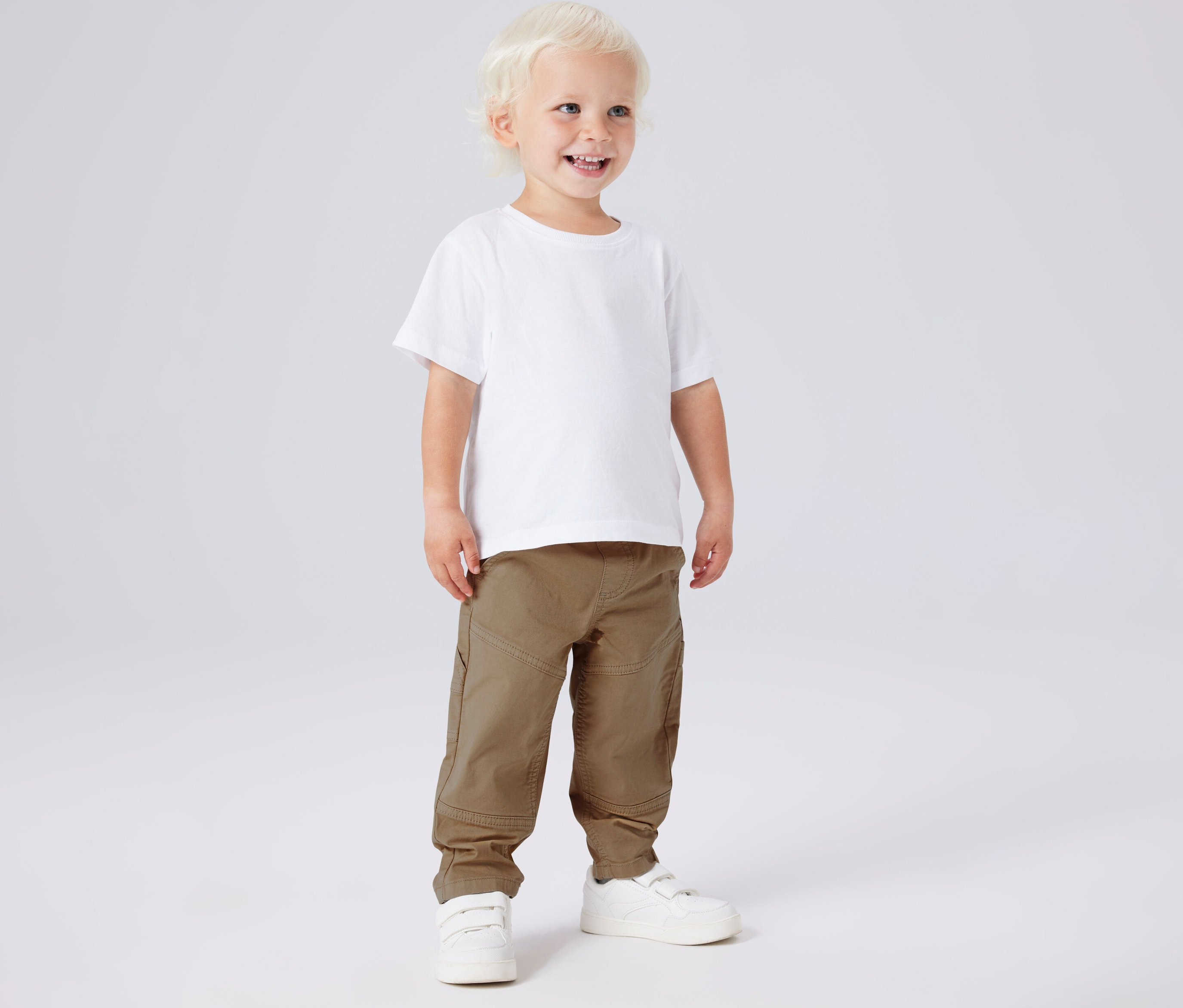 Uśmiechnięty chłopiec w białej koszulce i khaki spodniach z diagonalu NAME IT oraz białych butach.