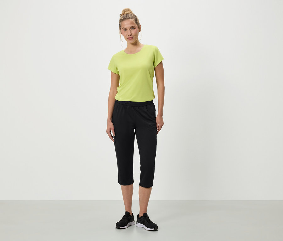 Kobieta stoi na białym tle w zielonej koszulce, czarnych spodniach rybaczkach i czarnych butach sportowych.