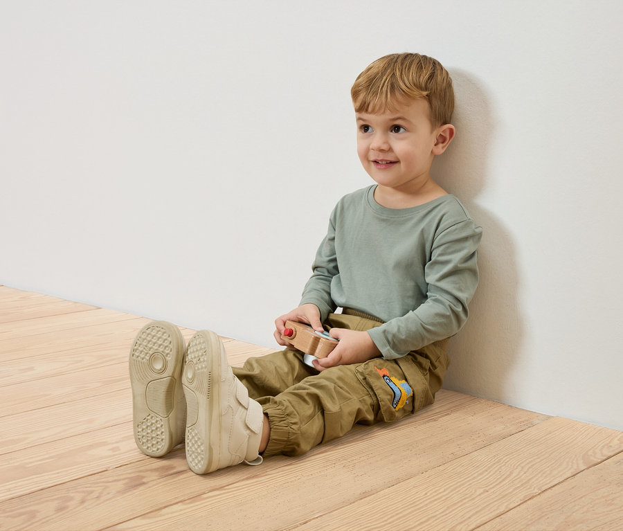 Uśmiechnięty chłopiec siedzi na drewnianej podłodze, oparty o białą ścianę, trzymając drewniany samochodzik.