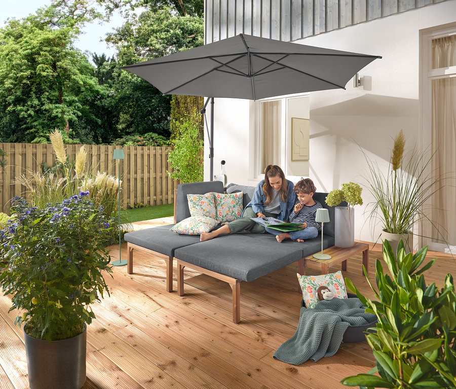 Kobieta i dziecko siedzą na szarej sofie ogrodowej z oddzielnie wysuwanymi podnóżkami. Na sofie leżą dwie poduszki ogrodowe we wzory kwiatowe. Antracytowy parasol Leco z podstawą i pokrowcem zapewnia cień. Na drewnianym stoliku obok sofy stoi szałwiowa lampa stołowa LED z akumulatorem, a w tle widać szałwiową lampę podłogową LED z akumulatorem.