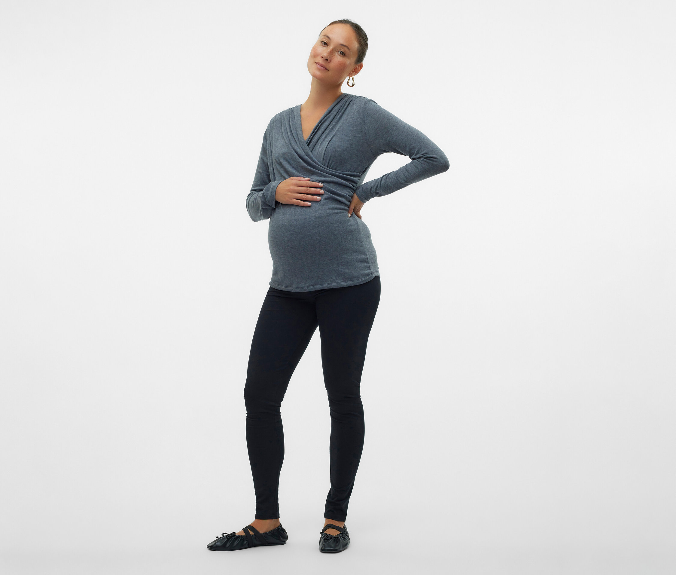 Kobieta w ciąży w szarym topie kopertowym, czarnych legginsach i balerinach pozuje na białym tle.