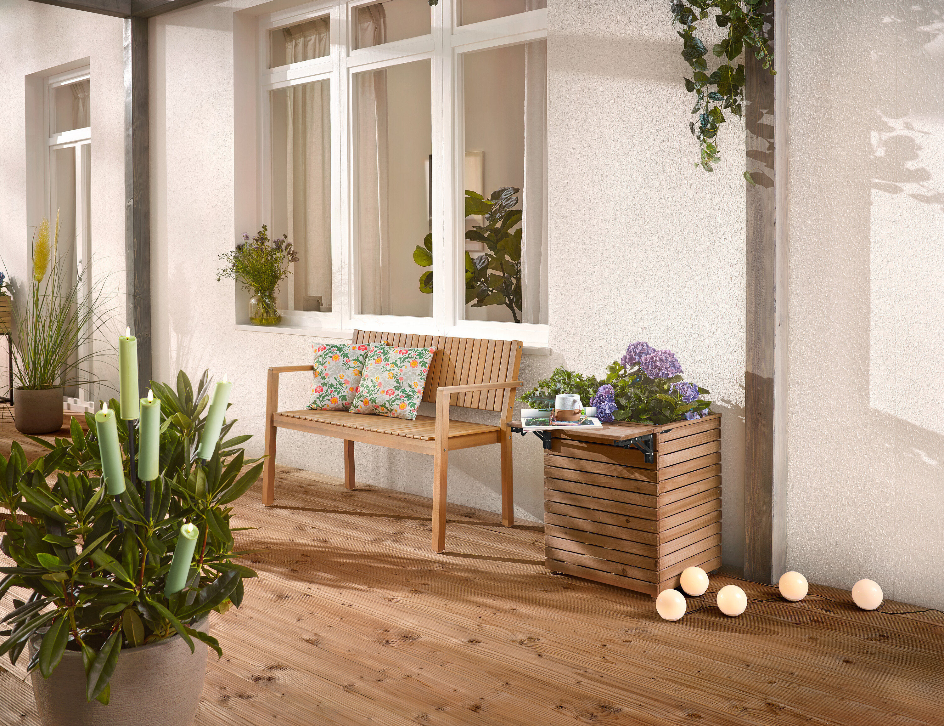 Na drewnianym tarasie stoi drewniana ławka z poduszkami, skrzynka na rośliny z kwiatami i roślina doniczkowa ze świecami.