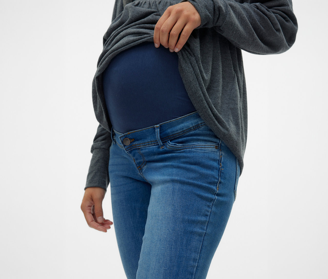 Kobieta podnosi szarą górę nad brzuchem, ma na sobie dżinsy skinny MAMALICIOUS „MLMILA” i granatową ciążową górę.