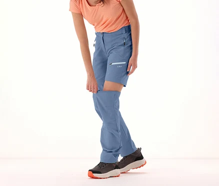 Kobieta odpina nogawki niebieskich spodni CMP z odpinanymi nogawkami. Nosi T-shirt i buty turystyczne.