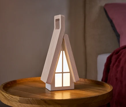 Lampa w kształcie domku z jasnymi drewnianymi bokami i świecącymi białymi oknami stoi na okrągłym drewnianym stoliku. W tle widać łóżko z różową poduszką i czerwoną narzutą.