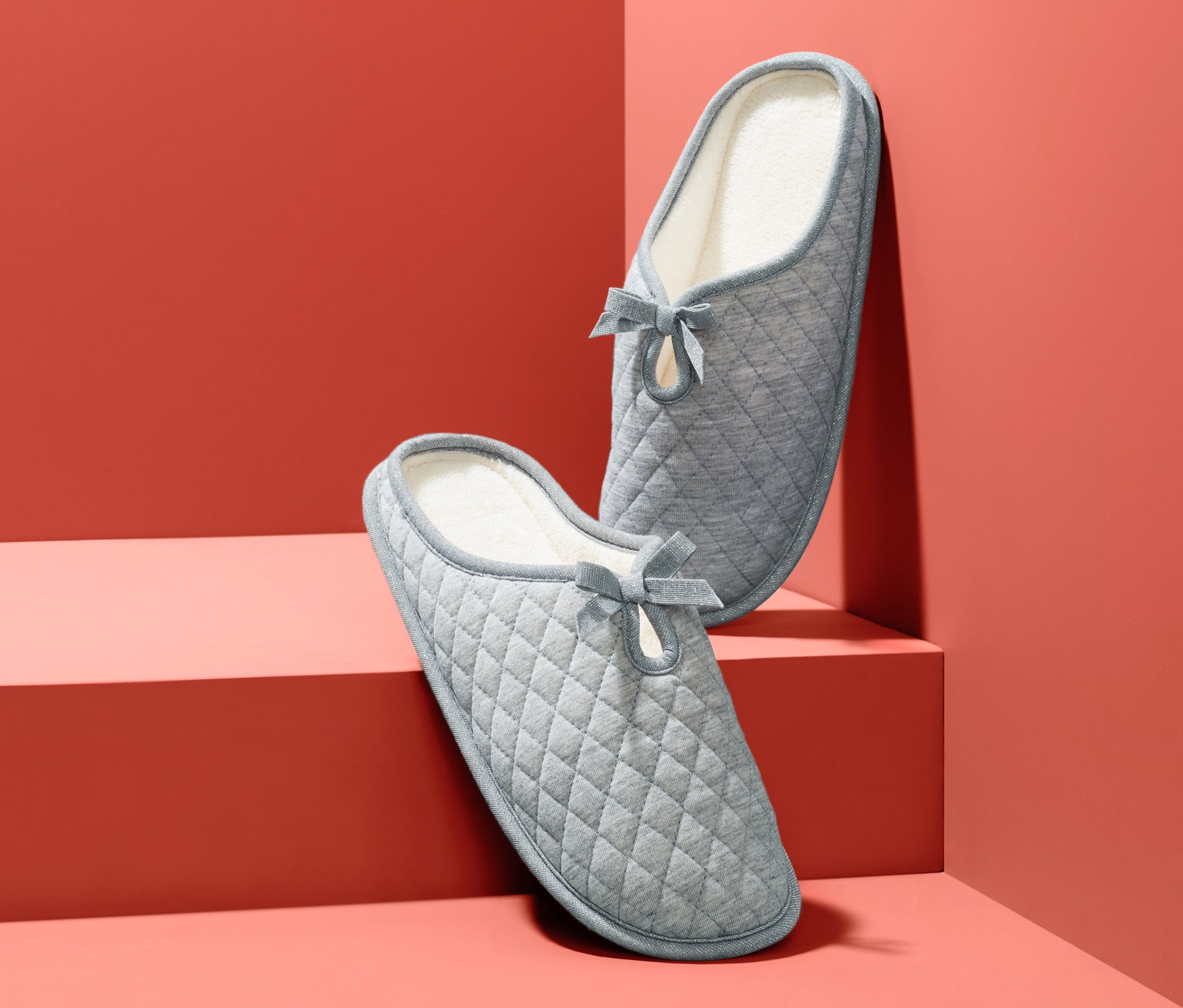 Dwa szare, pikowane kapcie z kokardą na koralowym tle.