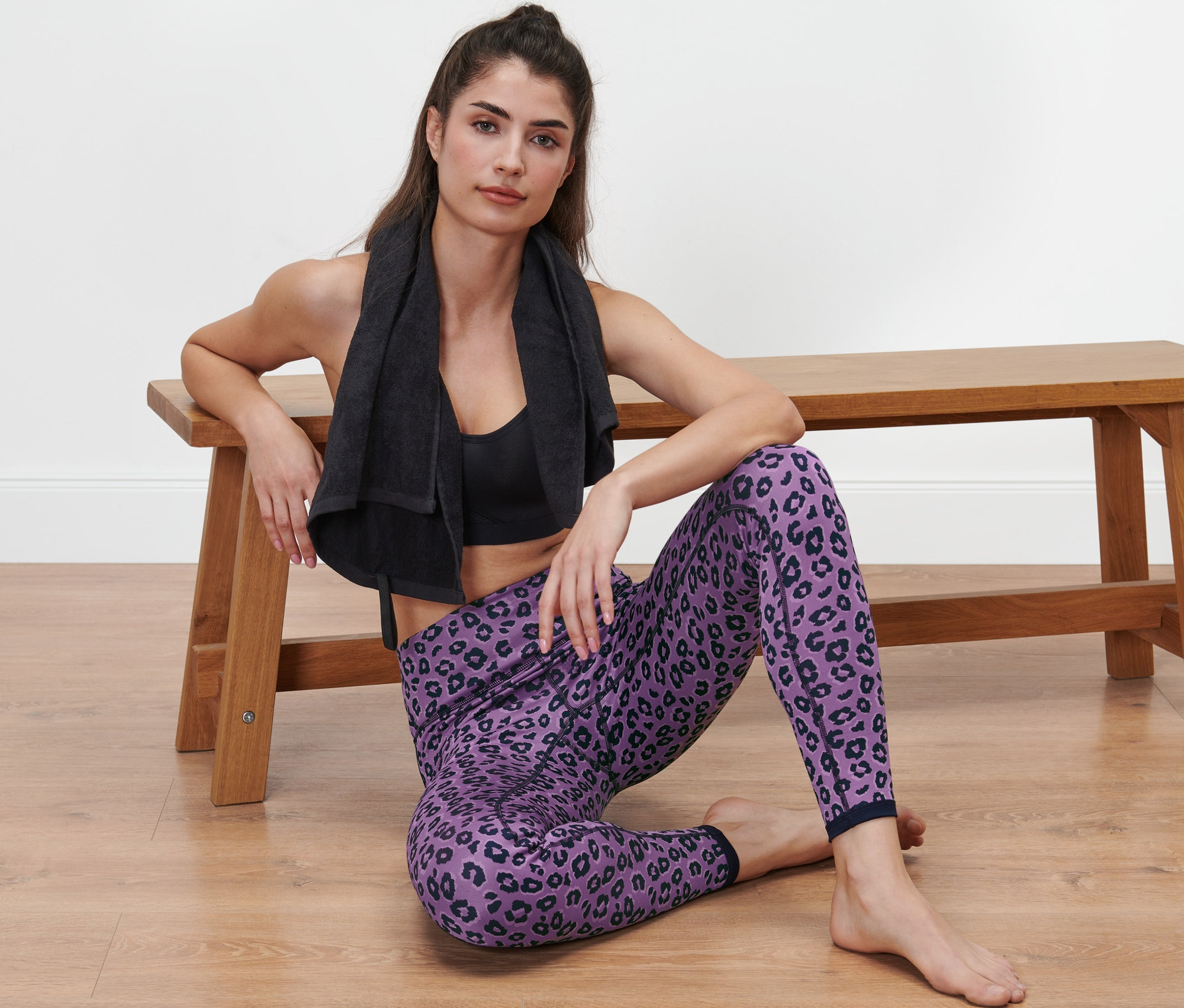 Kobieta siedzi na podłodze obok ławki, ubrana w dwustronne legginsy sportowe w kolorze niebieskim i lawendowym.