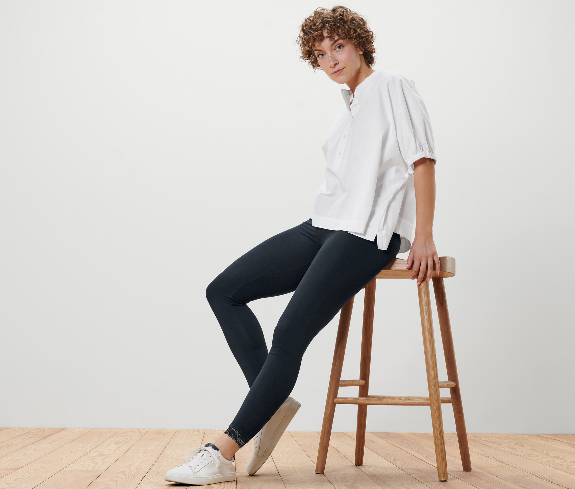 Kobieta siedzi na stołku, ubrana w czarne legginsy zakończone koronką i białą bluzkę.
