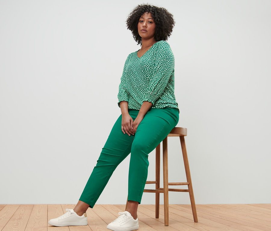 Kobieta siedzi na stołku ubrana w zieloną wzorzystą bluzkę i zielone spodnie stretchowe.