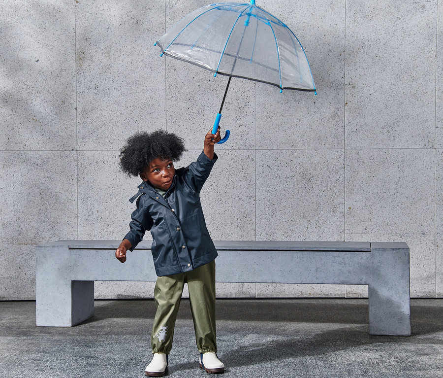 Chłopiec trzyma nad głową przezroczysty parasol z niebieskim obramowaniem i ma na sobie kurtkę przeciwdeszczową.