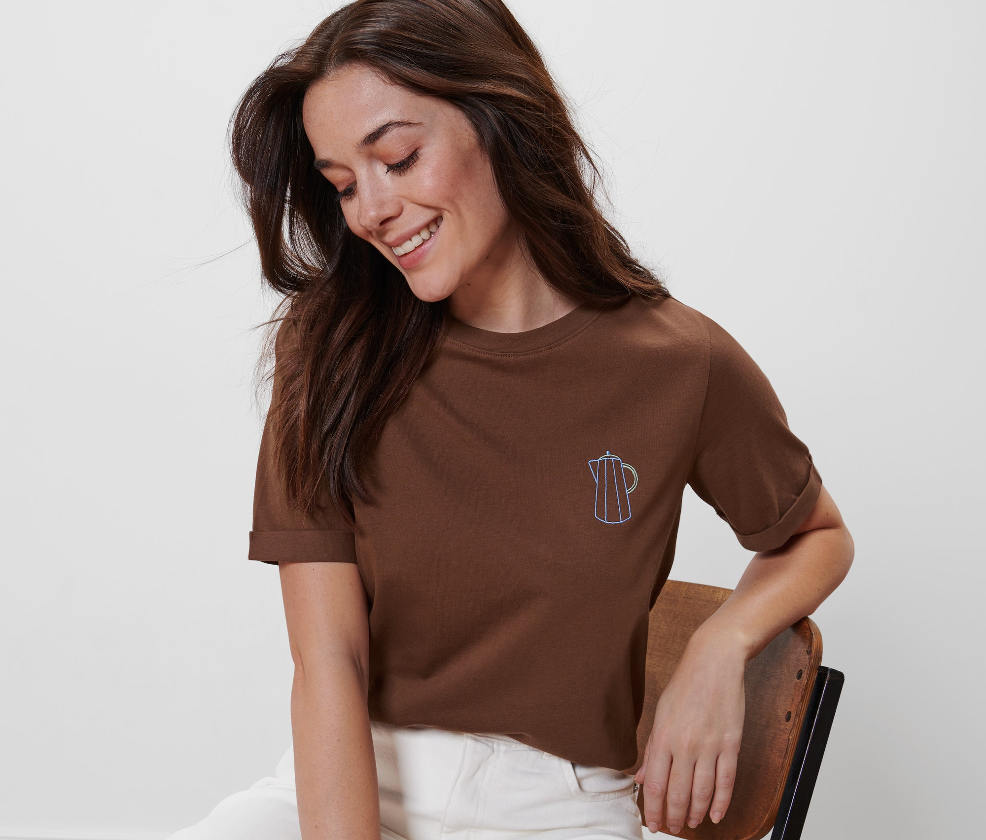 Kobieta siedzi na krześle i ma na sobie brązową koszulkę z haftem.