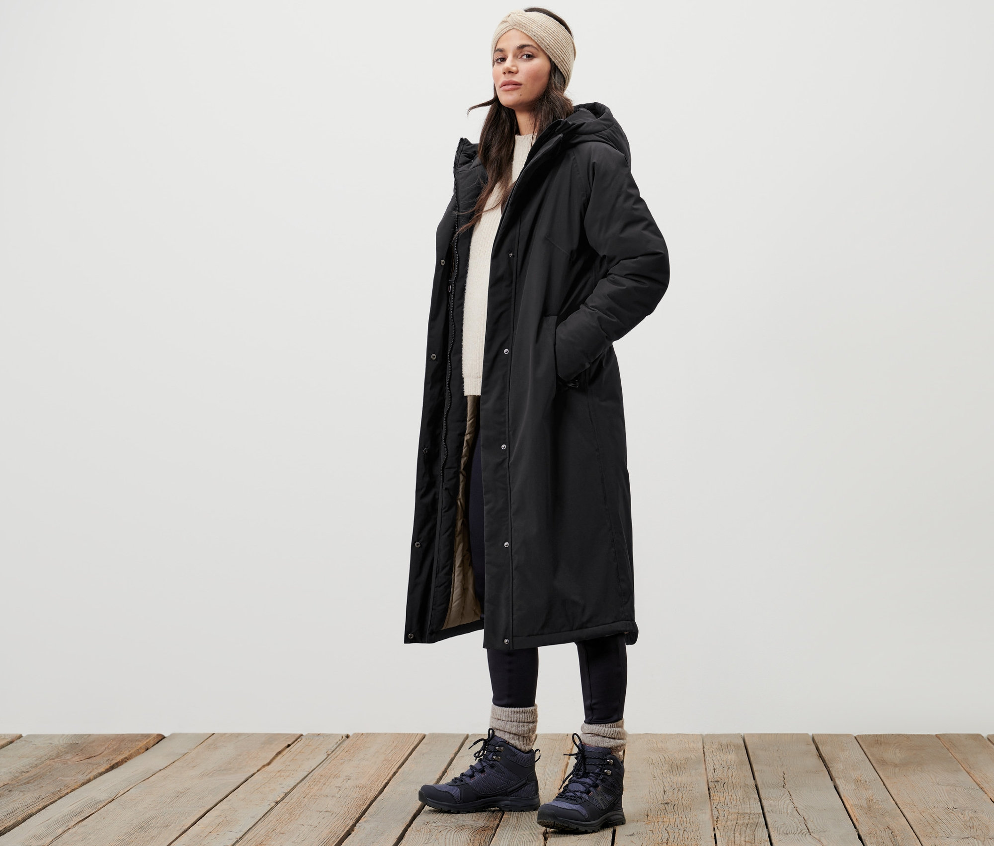 Kobieta stoi na drewnianej podłodze, ubrana w wyjątkowo długi płaszcz zimowy.