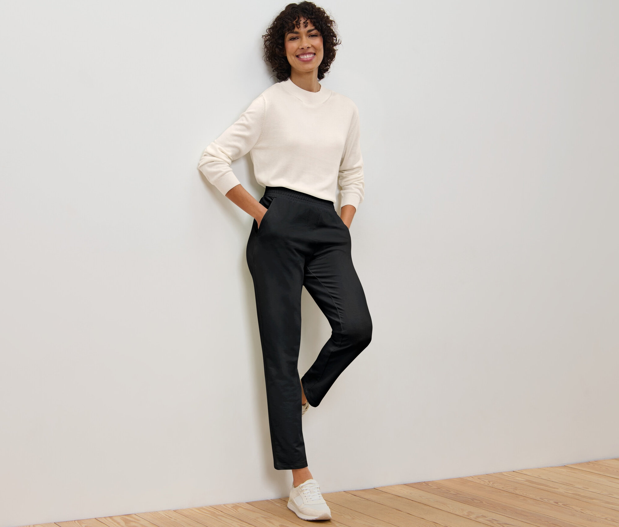 Kobieta oparta o ścianę ma na sobie czarne spodnie joggery i kremowy top.