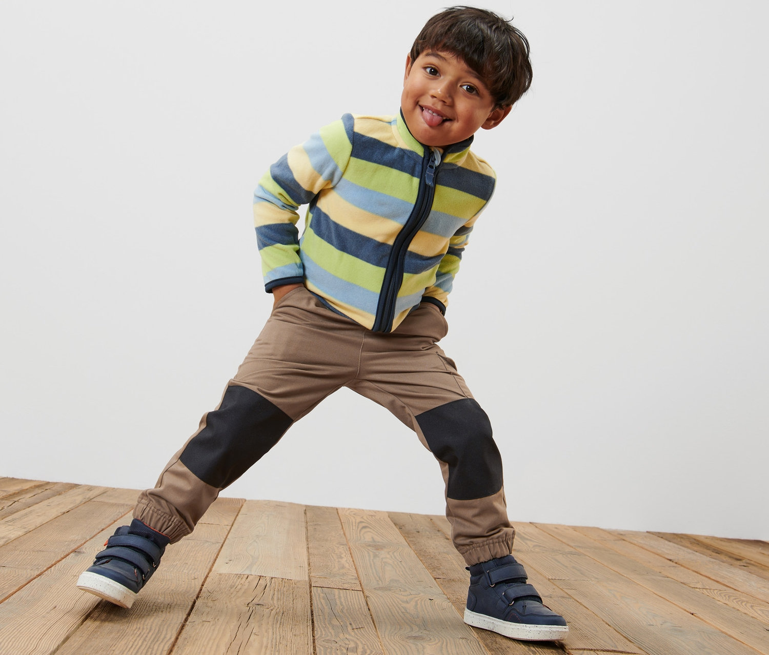 Chłopiec stoi na drewnianej podłodze w spodniach typu cargo na gumce, w bluzie w paski i butach.