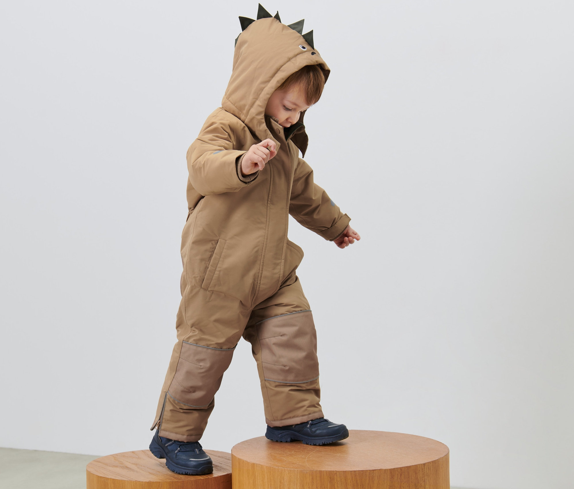 Dziecko w kombinezonie zimowym z kapturem w kształcie kolców stoi na drewnianych stołkach.