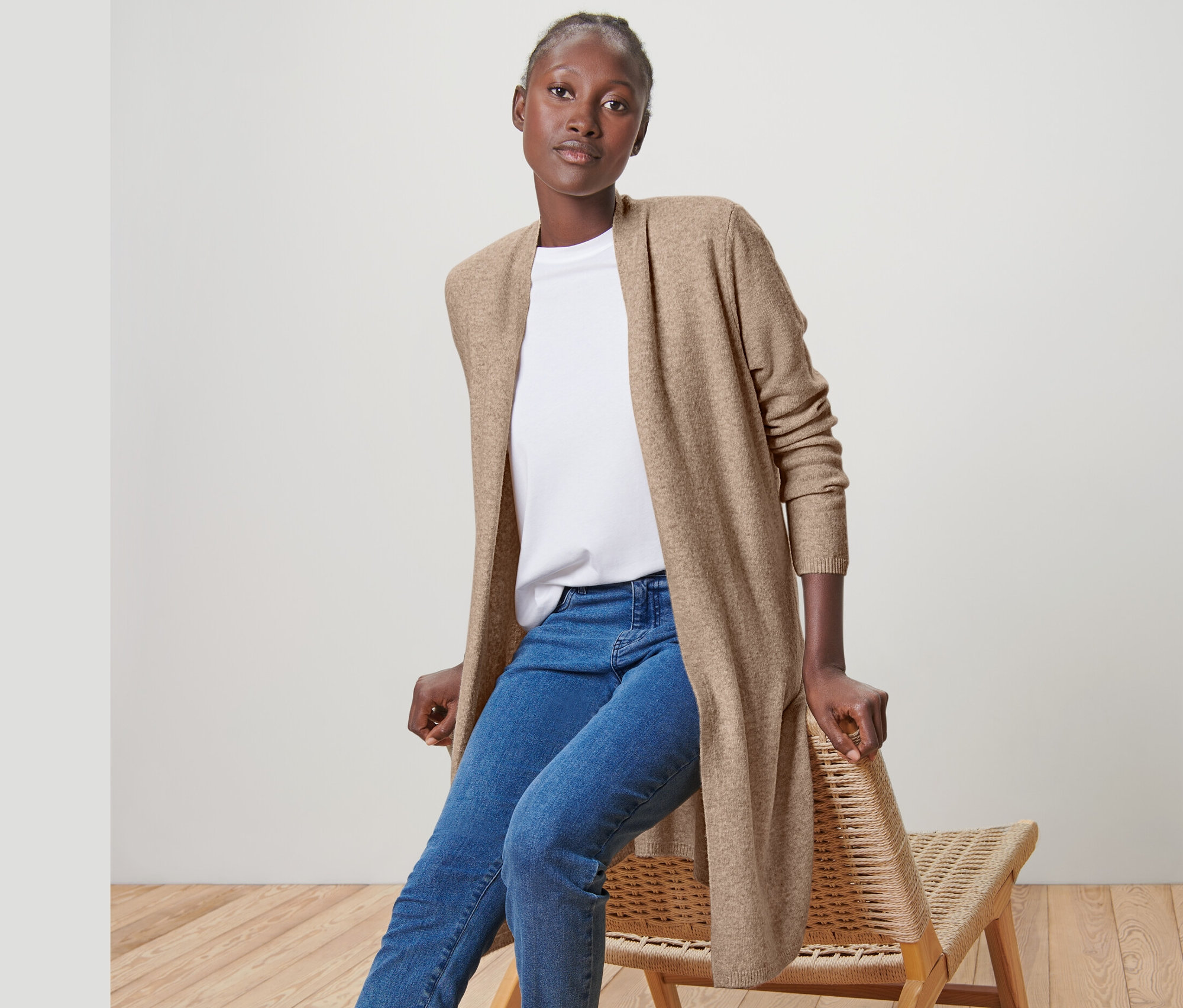 Kobieta siedzi na krześle i ma na sobie długi kardigan, dżinsy i tenisówki.