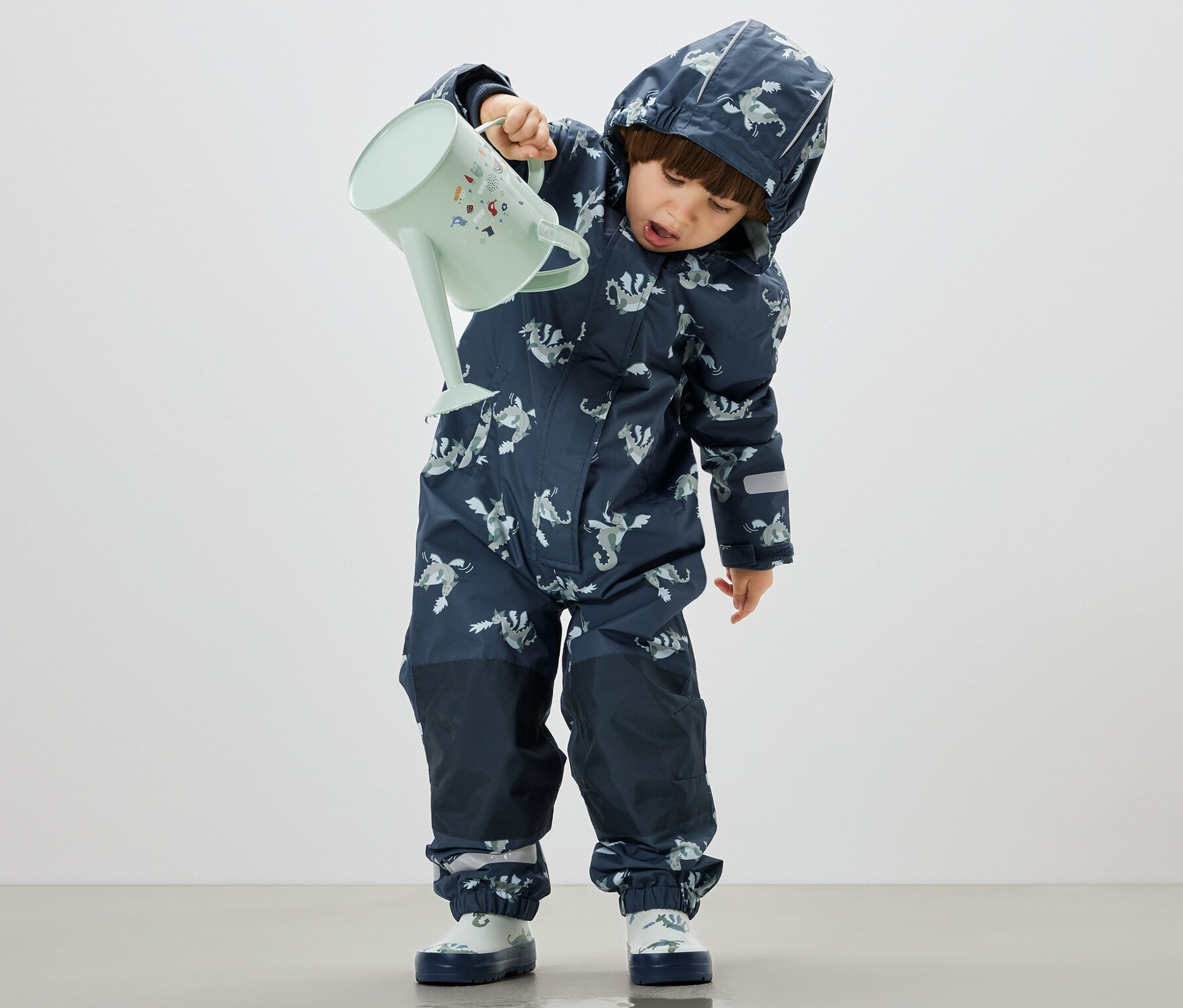 Chłopiec wylewa wodę z pastelowo zielonej konewki na podłogę, ubrany w Dziecięcy kombinezon przeciwdeszczowy, rycerz.