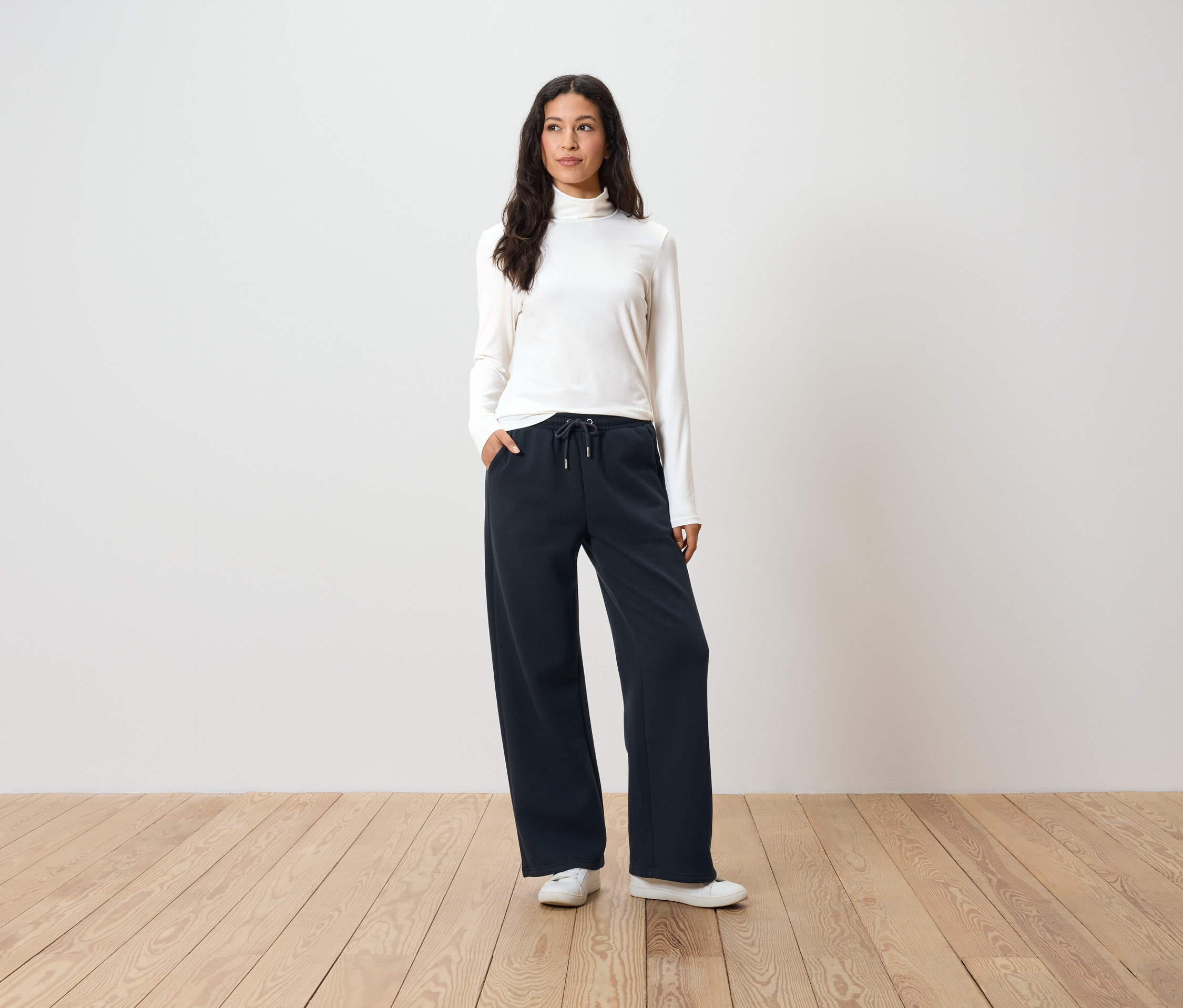 Kobieta stoi z rękami w kieszeniach granatowych, szerokich spodni, ma na sobie biały golf i białe tenisówki.