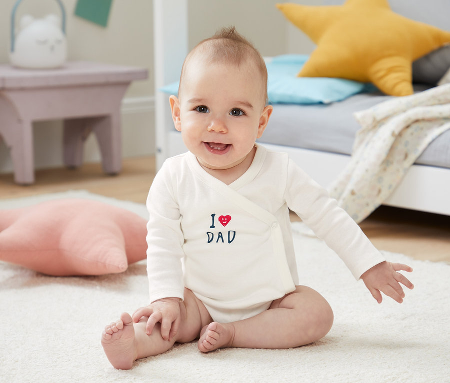 Uśmiechnięte dziecko siedzi na białym dywanie, ubrane w białe body z długim rękawem z bawełny ekologicznej z napisem "I love Dad".