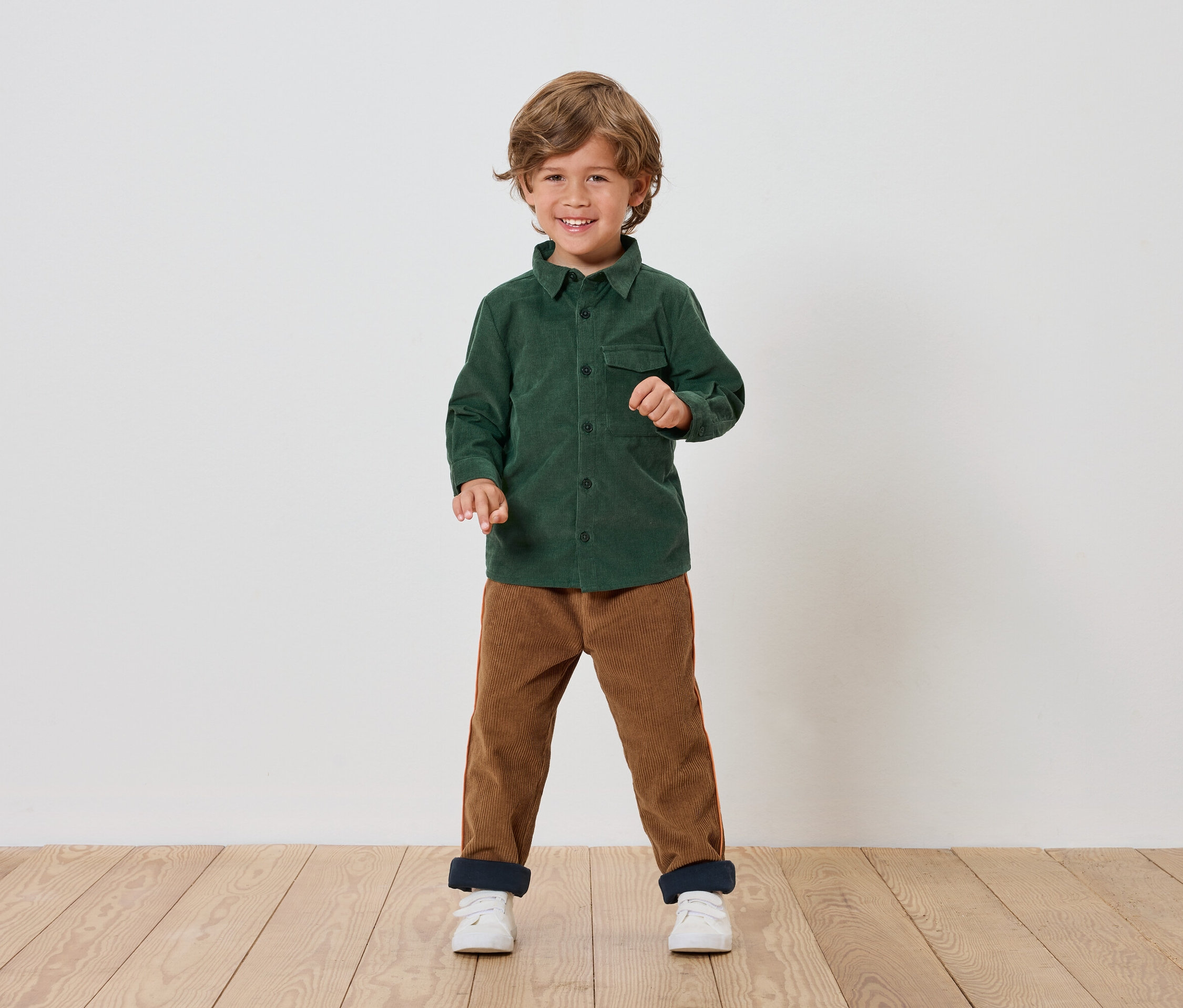 Chłopiec stoi i uśmiecha się, ubrany w zieloną koszulę, brązowe spodnie i białe buty.