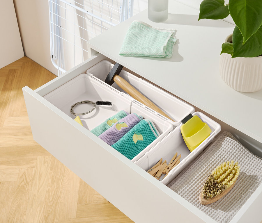 Otwarta szuflada białej szafki z różnymi przedmiotami, w tym ściereczkami, młotkiem i szczotką.