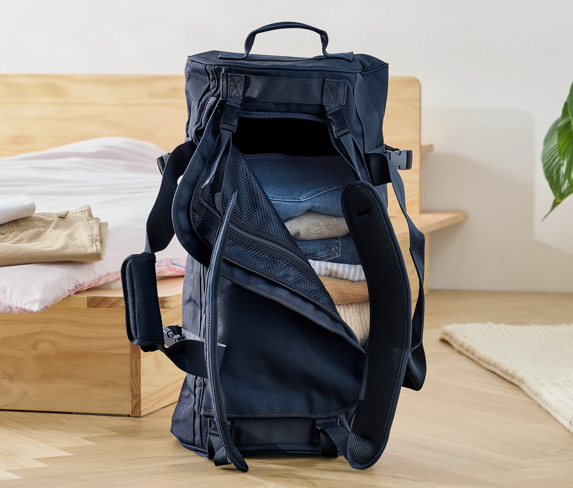 Otwarta torba i plecak w jednym wypełniona ubraniami stoi na podłodze obok łóżka.