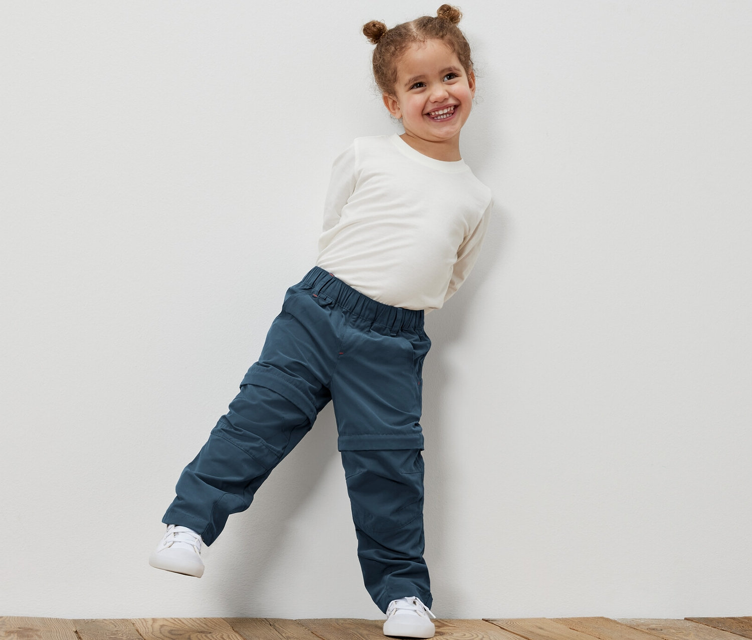 Uśmiechnięta dziewczynka opiera się o białą ścianę, ubrana w spodnie funkcyjne dla małych dzieci i białą koszulę.