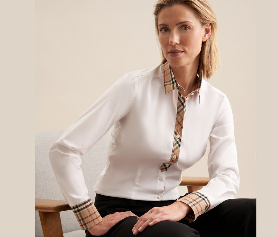 Kobieta siedzi na krześle i ma na sobie białą popelinową koszulę Seidensticker z detalami w kratę.