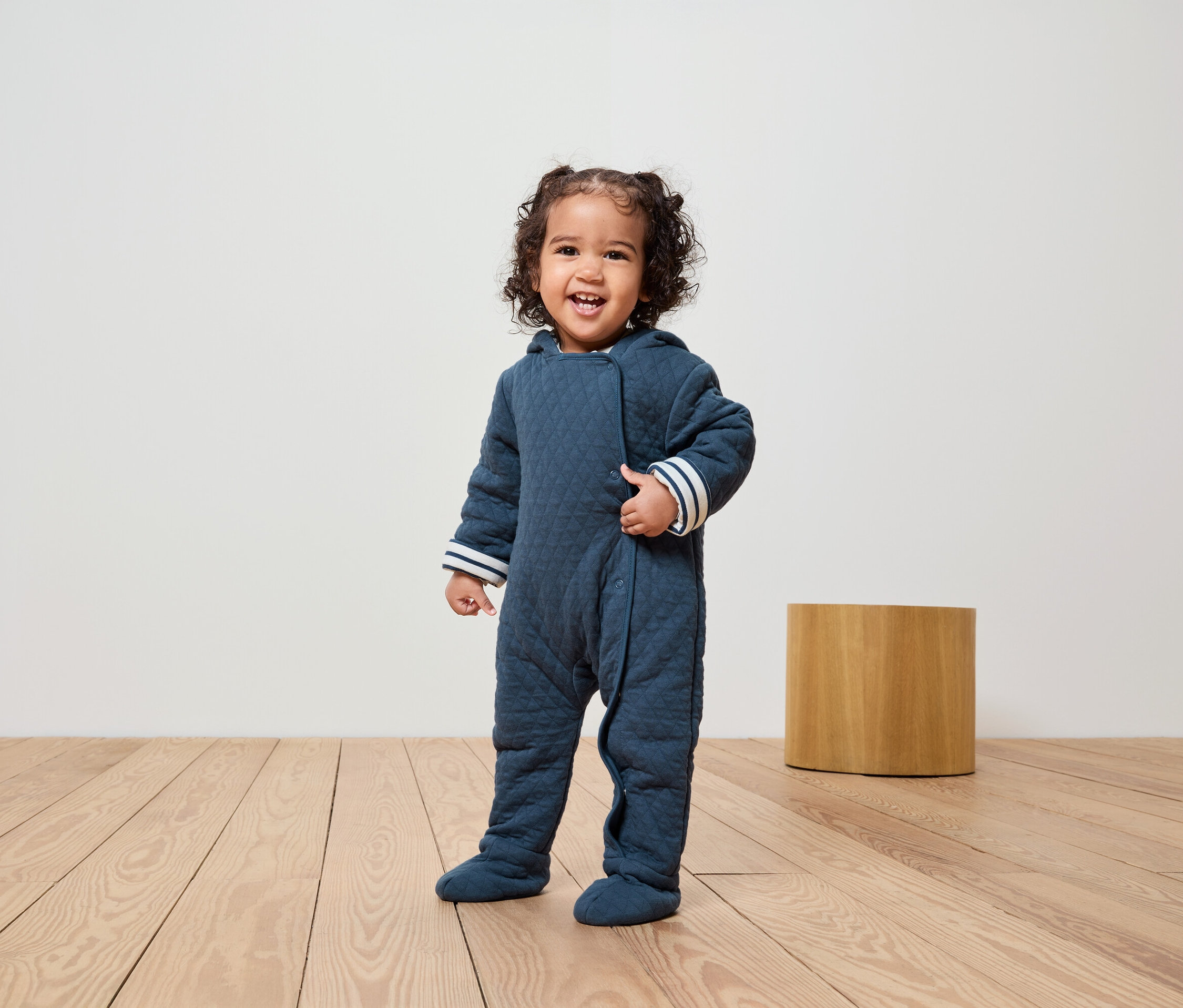 Uśmiechnięte dziecko stoi na drewnianej podłodze, ubrane w granatowy, pikowany kombinezon z kapturem i białymi paskami na mankietach.