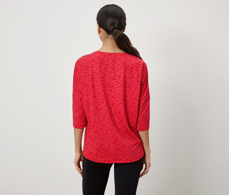 Z tyłu widać kobietę z włosami związanymi w koński ogon, ubraną w czerwoną bluzkę z rękawami trzy czwarte i zwierzęcym wzorem.