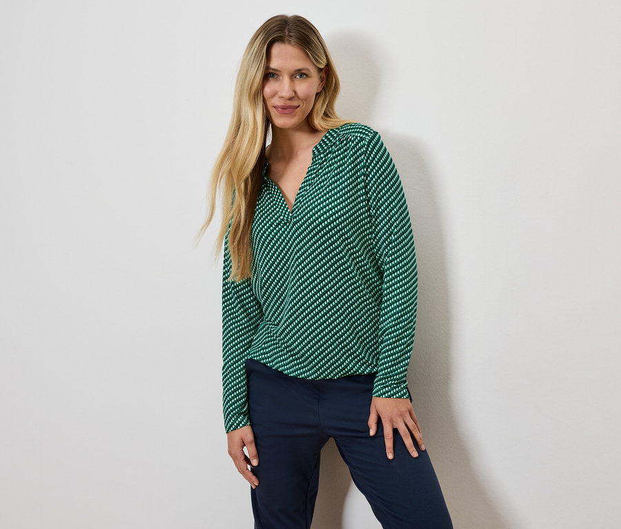 Kobieta o blond włosach pozuje w zielonej bluzce w paski i granatowych spodniach.