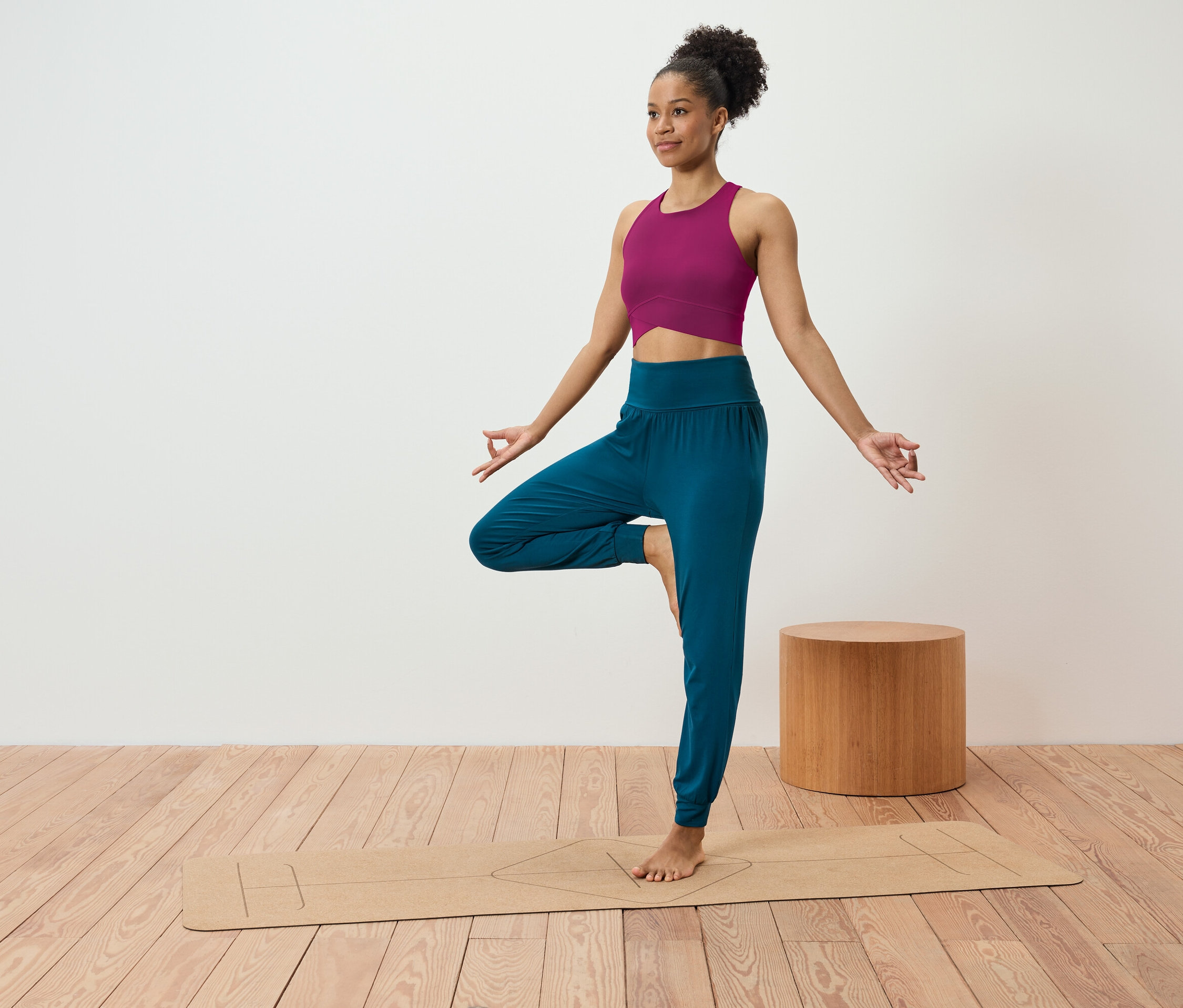 Kobieta w różowym topie i niebieskich spodniach stoi w pozycji drzewa na macie do jogi.