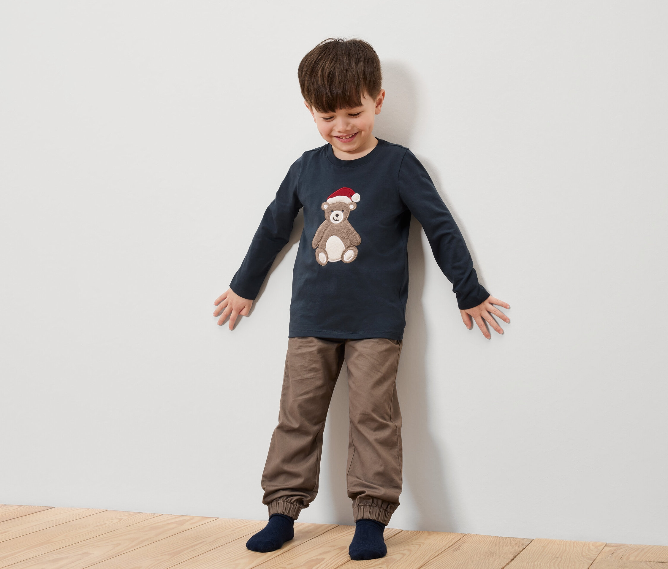 Chłopiec stoi przed białą ścianą, ubrany w granatową koszulę z misiem w czapce Świętego Mikołaja i brązowe spodnie.