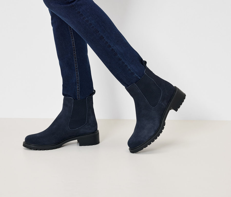 Nogi w granatowych dżinsach i granatowych zamszowych botkach Chelsea na niskim obcasie, idące po białej powierzchni.