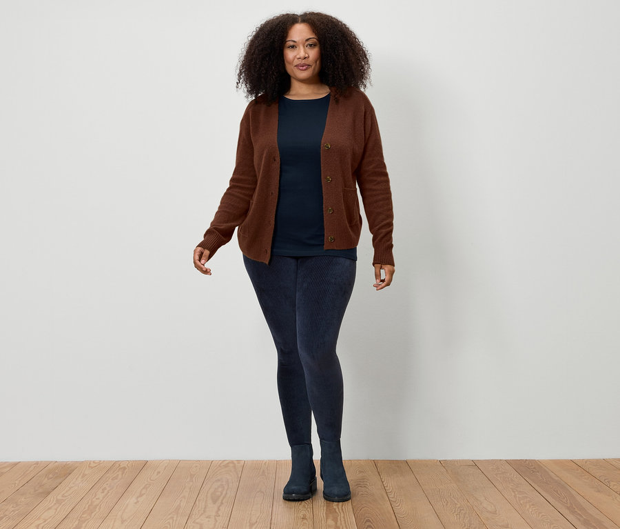 Kobieta z brązowymi kręconymi włosami stoi na tle białej ściany, ubrana w brązowy kardigan, granatowy t-shirt, granatowe legginsy i granatowe buty.