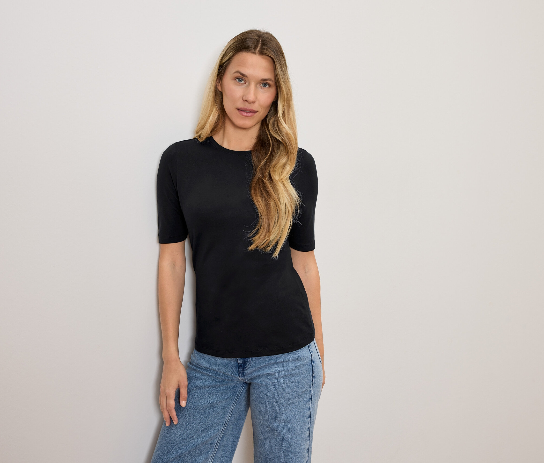 Kobieta o blond włosach pozuje w czarnej koszulce i niebieskich dżinsach.