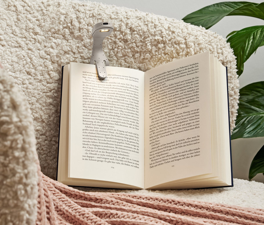 Szara lampka do czytania przypięta do otwartej książki leżącej na miękkim, kremowym kocu z różowymi akcentami.