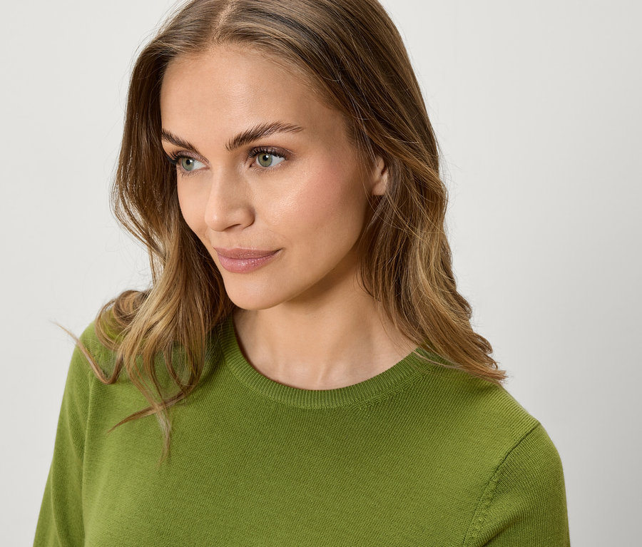 Portret kobiety z falowanymi, brązowymi włosami w zielonym swetrze, patrzącej w bok.