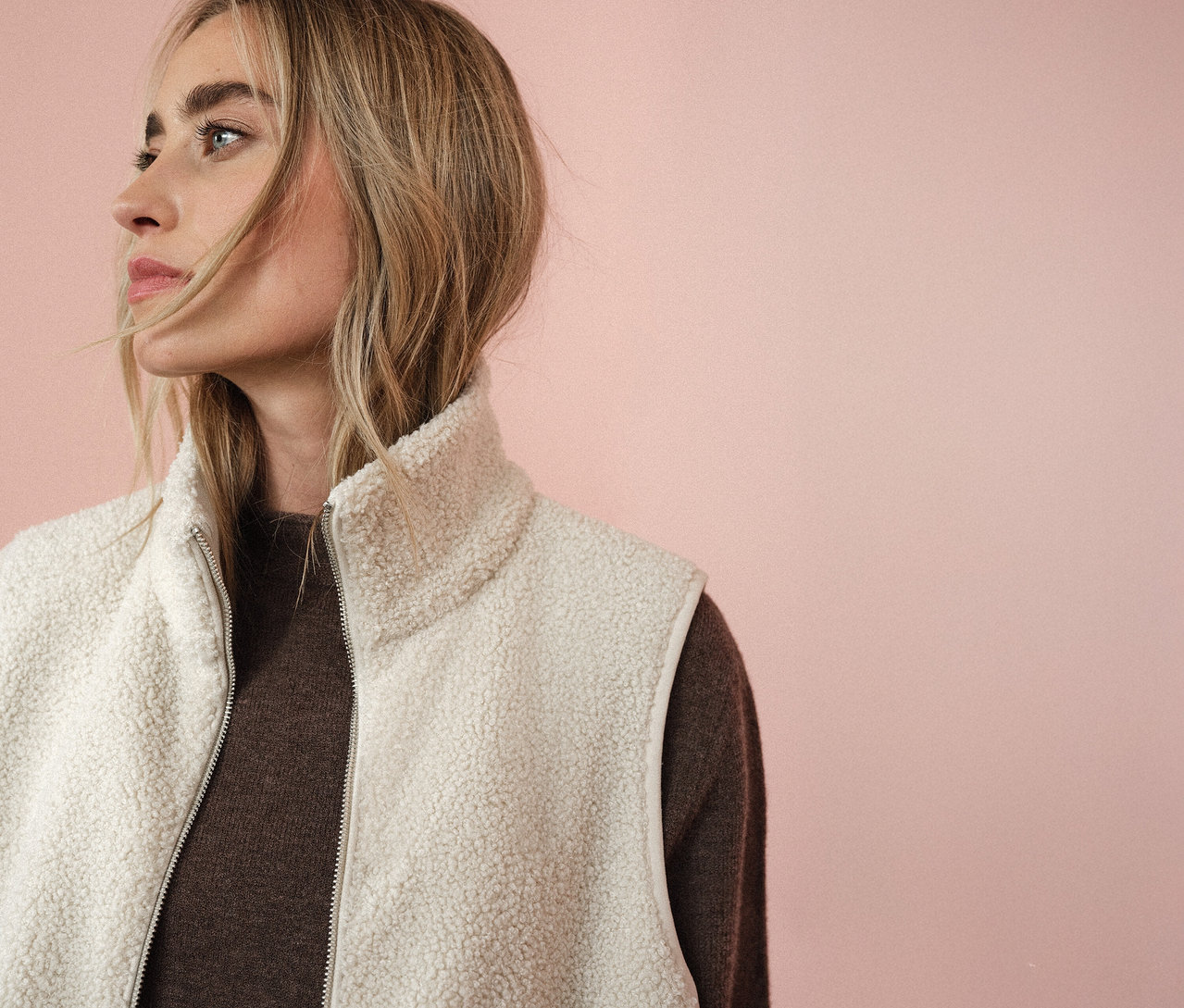 Kobieta w pluszowej kamizelce Street One na brązowym swetrze, patrzy w lewo na różowym tle.