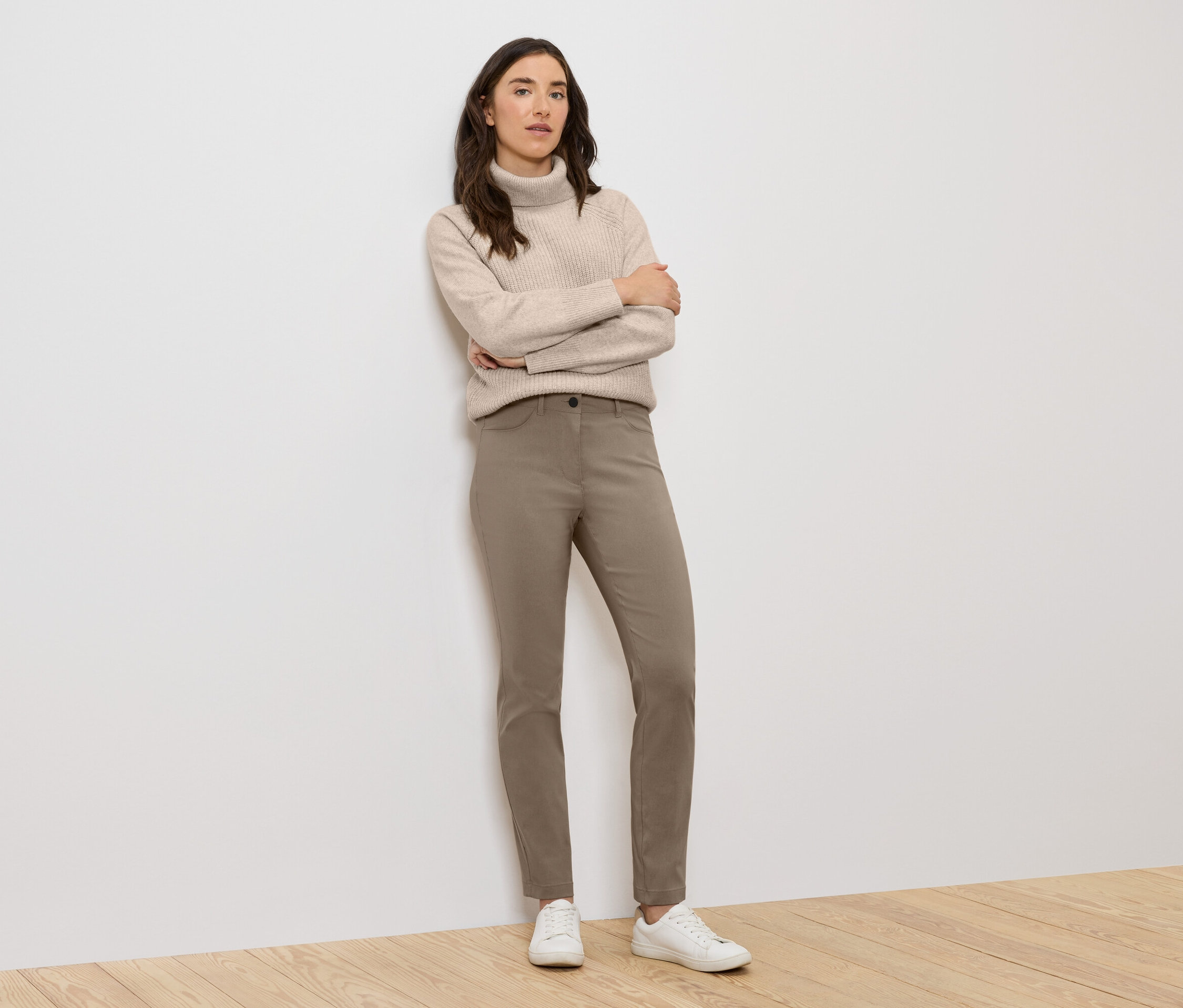 Kobieta stoi ze skrzyżowanymi ramionami, ubrana w beżowy golf, brązowe spodnie i białe tenisówki.