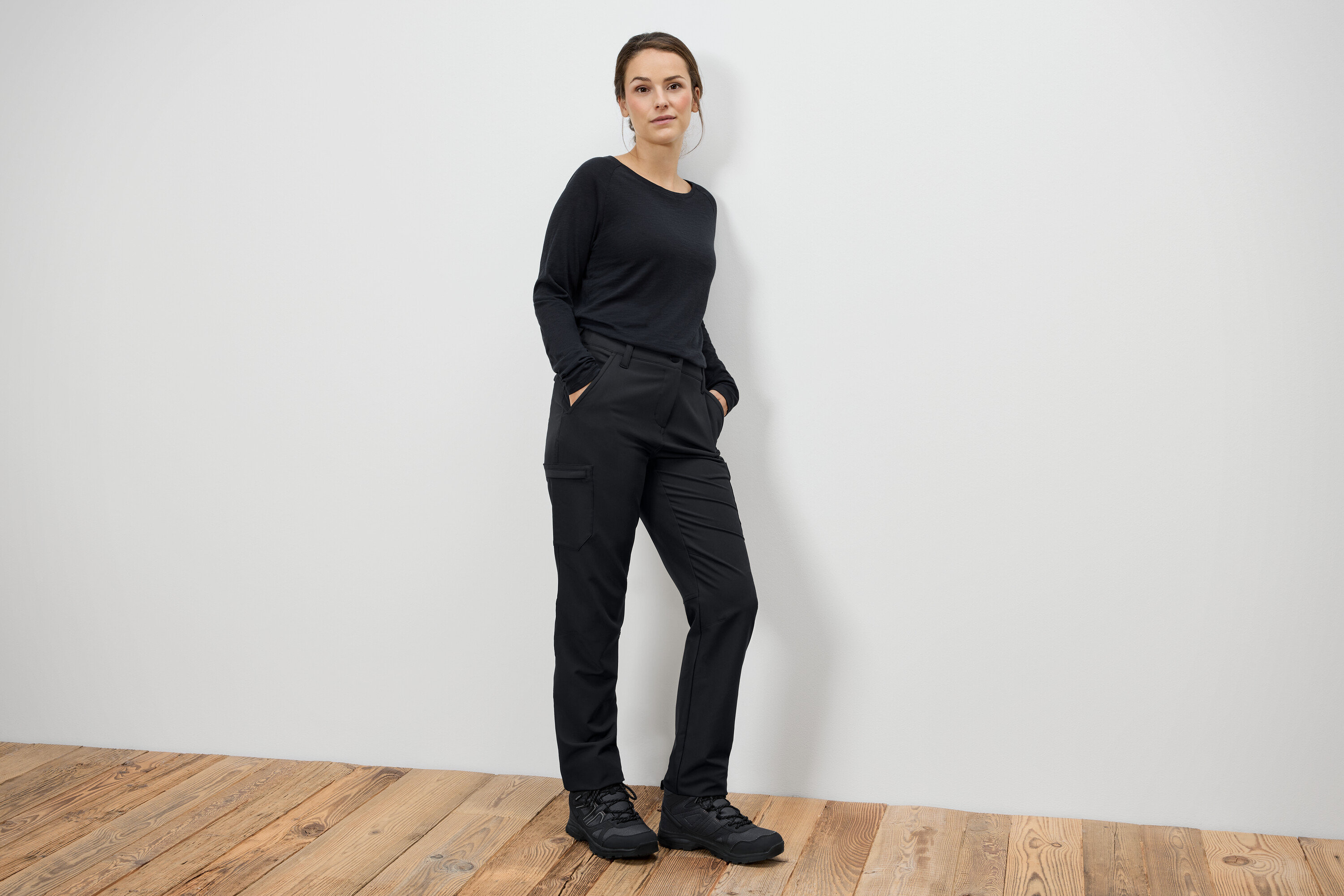 Kobieta stoi z rękami w kieszeniach w czarnych spodniach i czarnych butach.