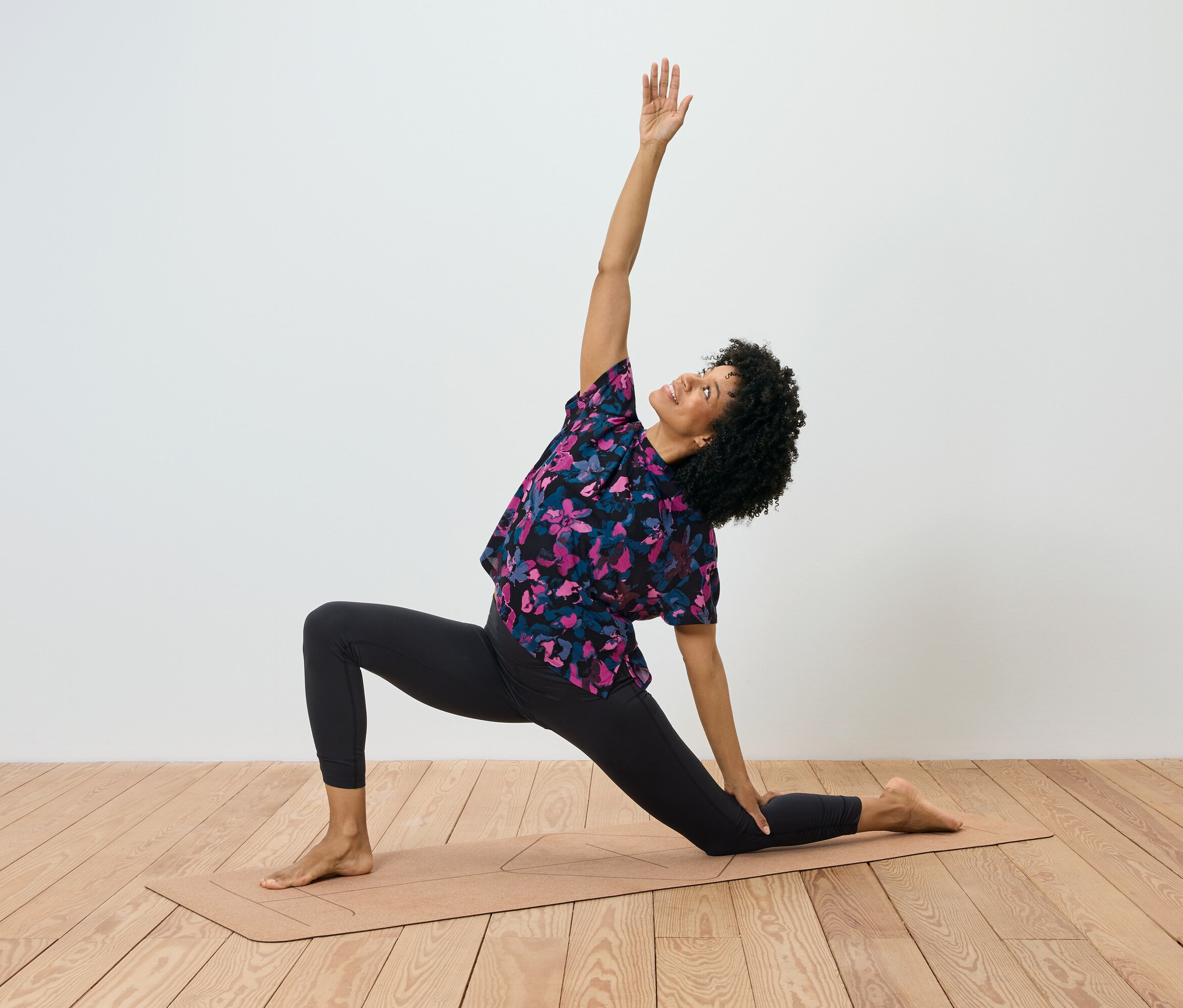 Kobieta z kręconymi włosami wykonuje ćwiczenie na macie do jogi, prawa ręka jest podniesiona, a lewa dotyka maty.