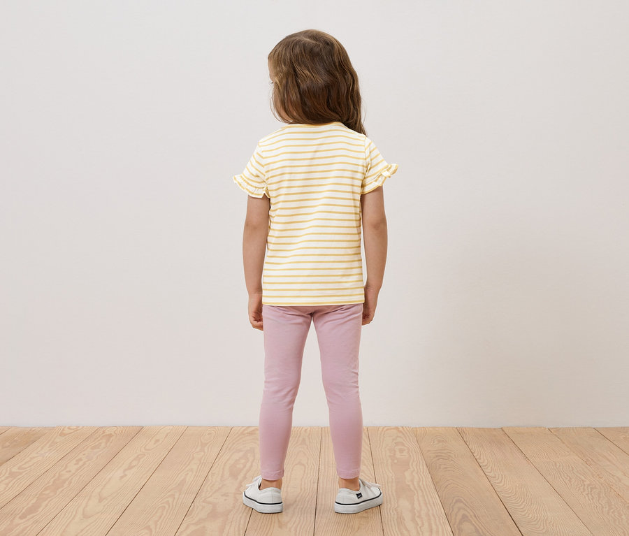Dziewczynka stoi tyłem, ma na sobie żółto-białą bluzkę w paski z falbankami, różowe legginsy i białe tenisówki.