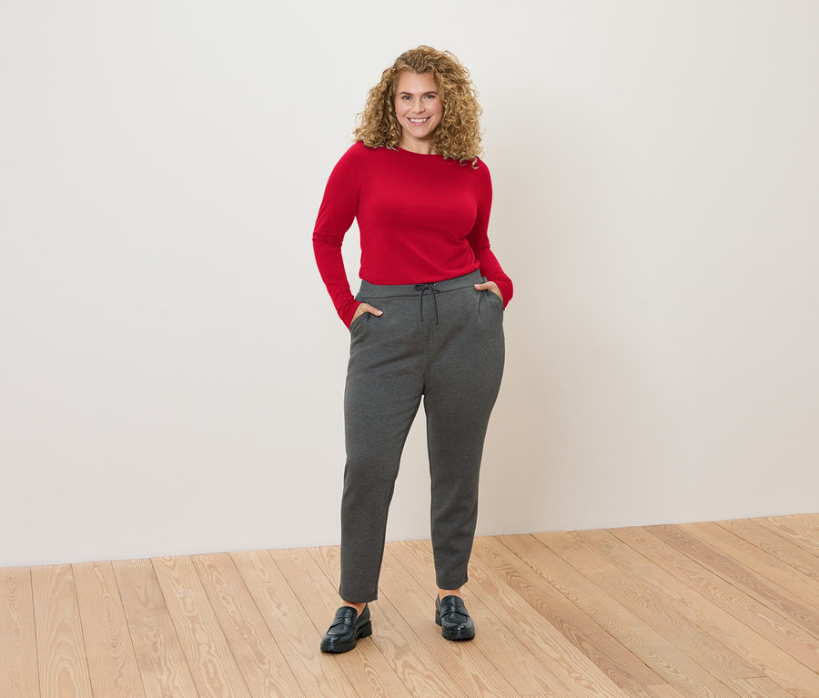 Kobieta z kręconymi włosami stoi na tle białej ściany, ubrana w czerwoną bluzkę z długim rękawem i szare spodnie dresowe.