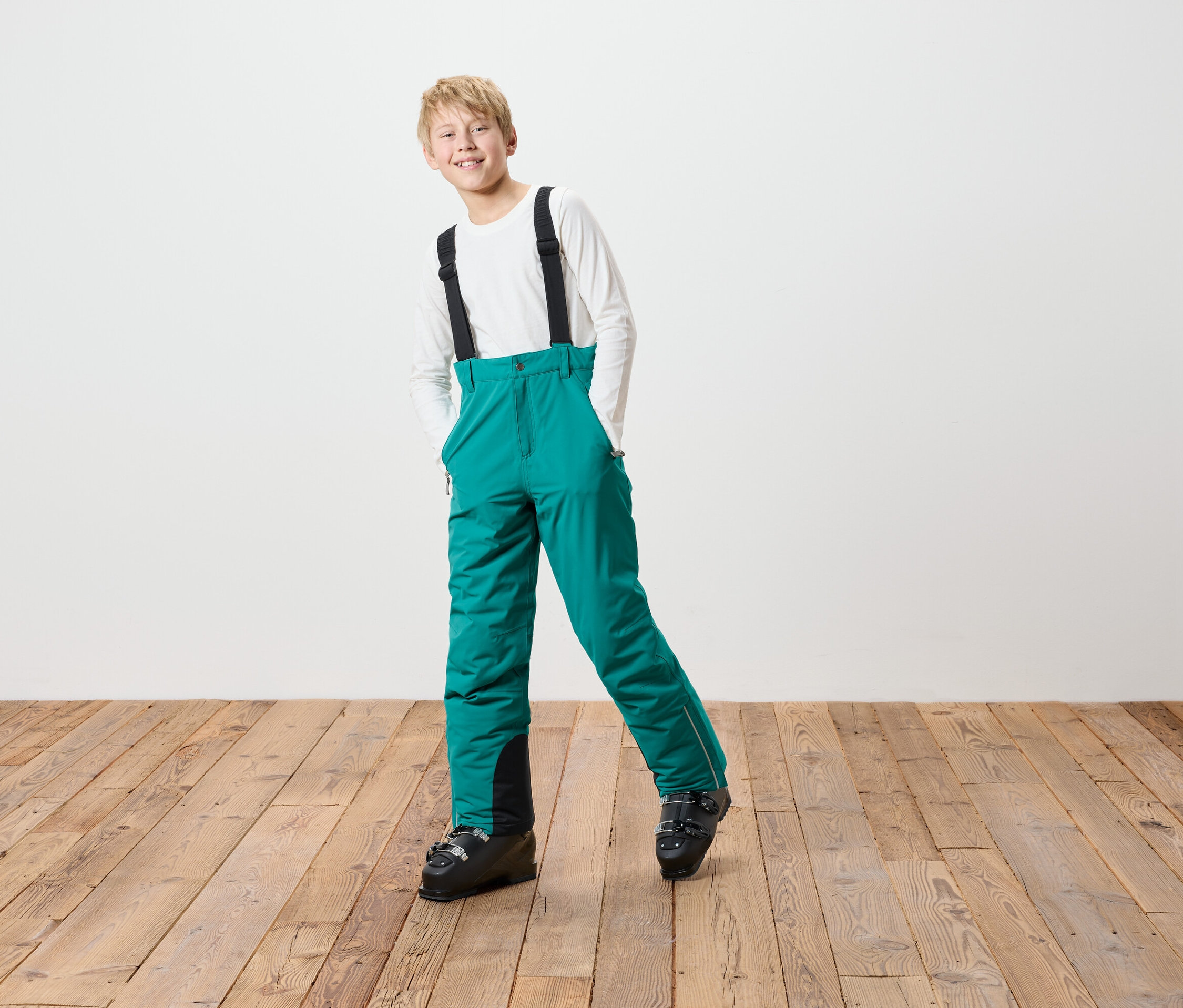 Chłopiec w zielonych spodniach narciarskich z czarnymi szelkami i butami narciarskimi stoi na drewnianej podłodze przed białą ścianą.