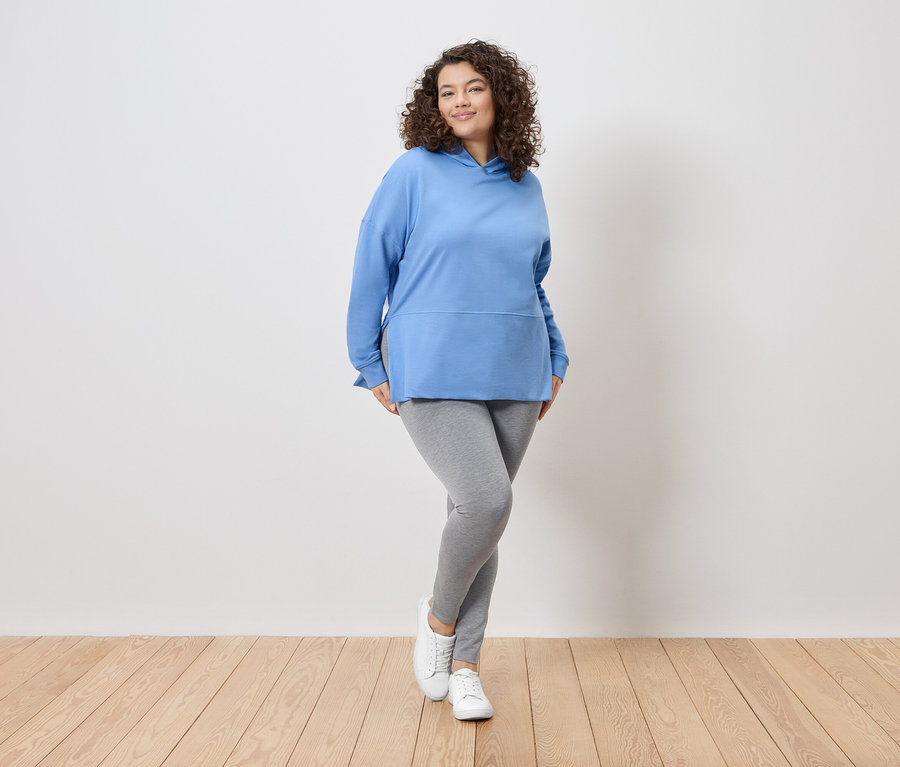 Kobieta z kręconymi włosami stoi na tle białej ściany, ubrana w niebieską bluzę z kapturem, szare legginsy i białe tenisówki.